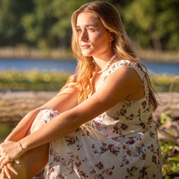 Golden-hour senior portrait of a young woman with long blonde hair in a floral dress, sitting on a lakeside log with calm water and trees in the background – natural senior photography by Imaginarium Pictures, West Michigan