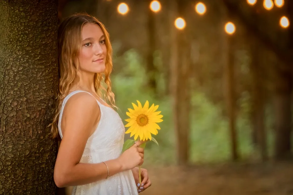 Natural senior portrait of a teenage girl with long wavy blonde hair in a white lace dress, leaning against a tree holding a bright sunflower, golden-hour string lights glowing in the wooded background – authentic senior photography by Imaginarium Pictures, West Michigan