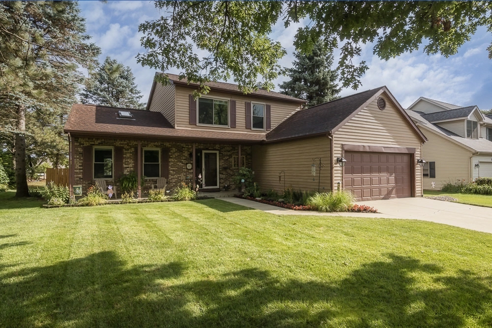 Exterior front view of a charming two-story brick and siding home in Portage, Michigan – well-maintained lawn, mature trees, welcoming porch with brick columns and flower beds – professional real estate photography by Imaginarium Pictures